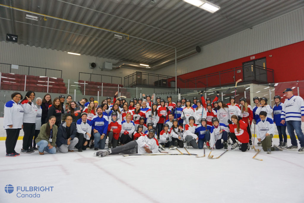 A large group of hockey players, students, and spectators on an ice rink all smiling for the camera!